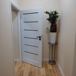 Sławomir Stańkowsk Usługi Remontowo-Wykończeniowe - Otwarta, biała, nowoczesna drzwi wewnętrzne z poziomymi, czarnymi paskami, widok na drewnianą podłogę, obok doniczka z rośliną na metalowym stojaku.