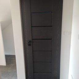 Sławomir Stańkowsk Usługi Remontowo-Wykończeniowe - Zamknięte, grafitowe drzwi wewnętrzne z poziomymi przeszkleniami, czarna klamka, białe ściany i szara podłoga z płytek.