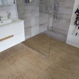 Nowoczesna łazienka z kabiną prysznicową typu walk-in, wykończona płytkami imitującymi drewno na podłodze i ścianach prysznica, biała szafka pod umywalką z drewnianym akcentem, oraz szklana ściana...