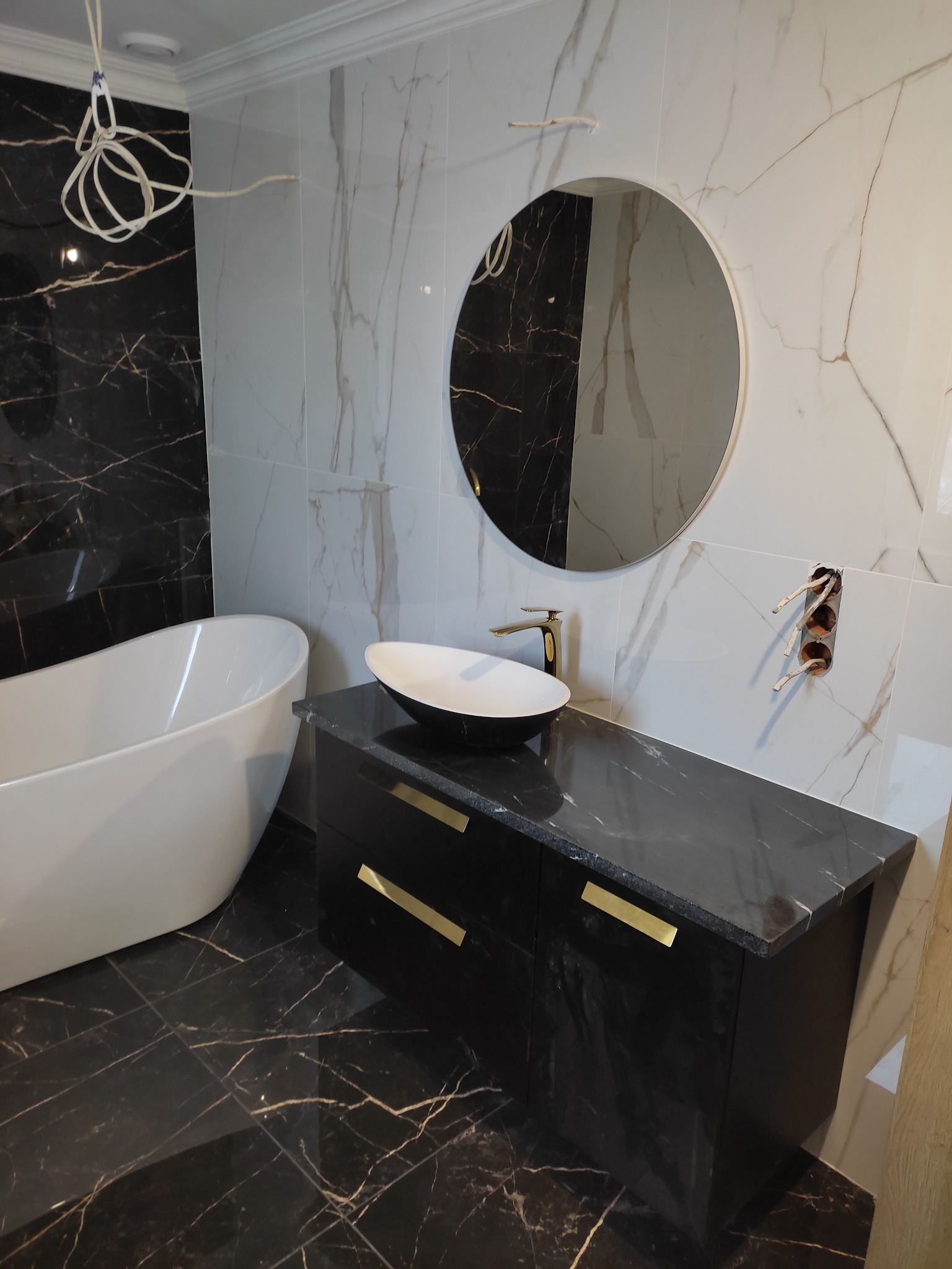 Nowoczesna łazienka w trakcie wykańczania: biała wanna wolnostojąca, czarna szafka z marmurowym blatem i złotymi uchwytami, okrągłe lustro, biało-czarne marmurowe płytki na ścianach i podłodze...