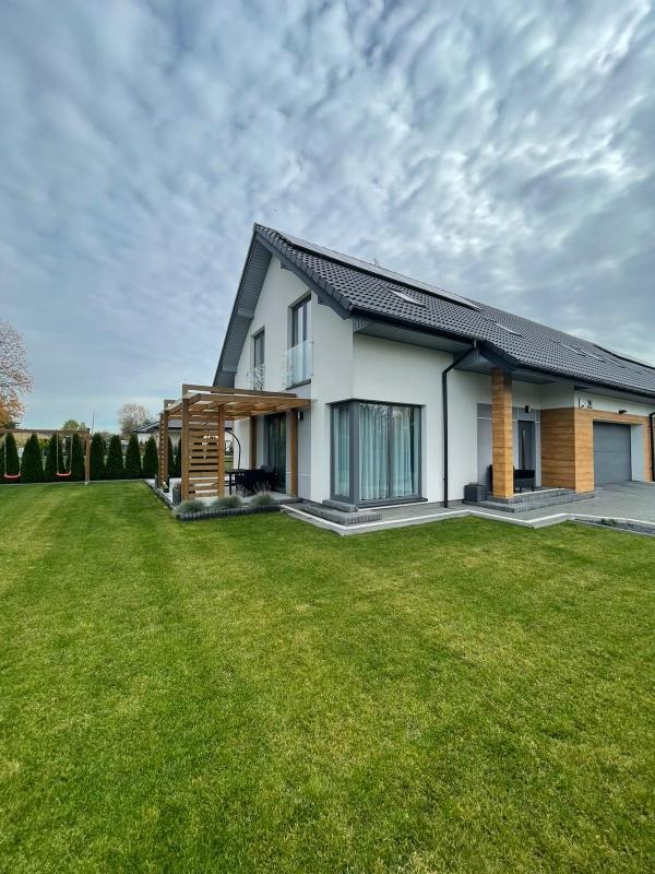 Nowoczesny dom jednorodzinny z płaskim dachem pokrytym czarną dachówką, drewnianą pergolą nad tarasem i starannie utrzymanym trawnikiem, uchwycony pod pochmurnym niebem.