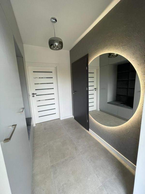 Nowoczesny przedpokój z szarą zabudową, podświetlanym okrągłym lustrem, białymi drzwiami z poziomymi frezami i szarą podłogą z płytek.