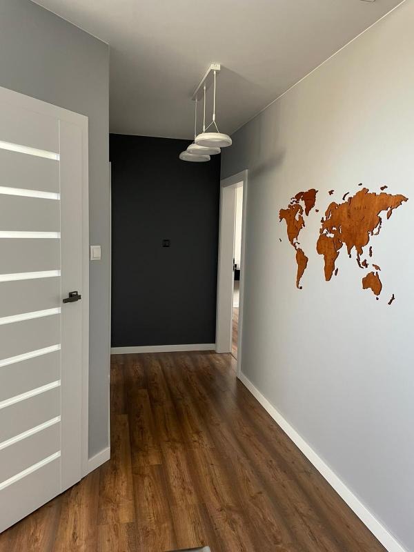 Nowoczesny korytarz z drewnianą mapą świata na szarej ścianie, podłoga z paneli imitujących drewno, białe drzwi z szarymi wstawkami i minimalistyczna lampa sufitowa.