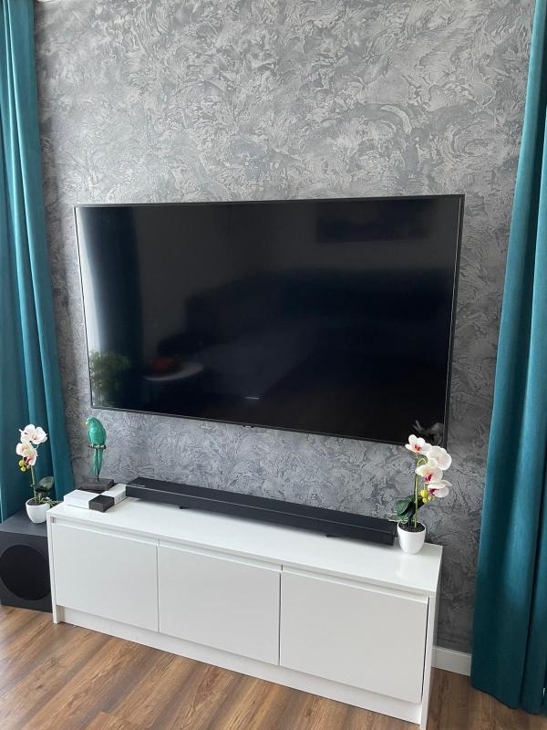 Telewizor na ścianie z dekoracyjną szarą tapetą, pod nim biała szafka RTV z głośnikiem soundbar, sztuczne kwiaty i turkusowe zasłony po bokach.