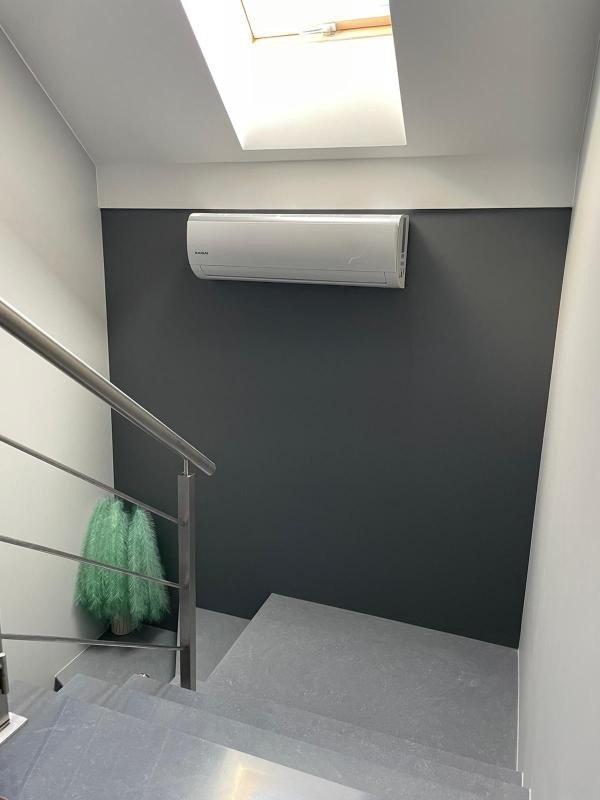 Klimatyzator zamontowany na ciemnoszarej ścianie nad schodami w Lublinie, widoczne schody wyłożone szarymi płytkami, metalowa barierka i nietypowa zielona dekoracja w rogu, oświetlenie naturalne...