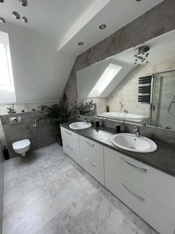 Nowoczesna łazienka na poddaszu z dwoma umywalkami, białymi szafkami z szarym blatem, dużym lustrem odbijającym wannę i oknem dachowym, szara płytka podłogowa i ścienna, toaleta w rogu.