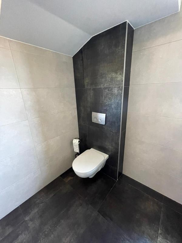 Nowoczesna łazienka z podwieszaną toaletą, szare płytki na ścianach i podłodze, ciemna ściana z ukrytym systemem spłukiwania, minimalistyczny design.