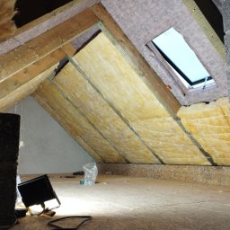 SkillWorks Michał Rzeszutek - Ocieplenie poddasza wełną mineralną, widoczne elementy konstrukcji drewnianej, okno dachowe, fragment ściany z bloczków betonowych oraz lampa robocza.