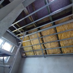 SkillWorks Michał Rzeszutek - Montaż izolacji z wełny mineralnej w metalowym stelażu pod płytę gipsowo-kartonową na poddaszu, widoczny fragment okna dachowego i rusztowania.