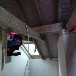 SkillWorks Michał Rzeszutek - Laser krzyżowy Bosch ustawiony na drewnianej belce, wyznaczający poziom na ścianie poddasza w trakcie prac wykończeniowych.
