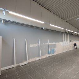 SkillWorks Michał Rzeszutek - Przygotowanie ściany w dużym pomieszczeniu do montażu regałów: szpachlowanie i malowanie na szaro, widoczne białe wsporniki, osoba w tle z wózkiem.