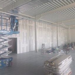 SkillWorks Michał Rzeszutek - Wnętrze hali w trakcie prac wykończeniowych, widoczne płyty kartonowo-gipsowe na ścianach przed szpachlowaniem, podnośnik koszowy po lewej stronie oraz materiały budowlane na podłodze.