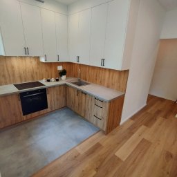 SkillWorks Michał Rzeszutek - Nowoczesna kuchnia z białymi szafkami górnymi i drewnopodobnymi szafkami dolnymi, szarym blatem i płytkami podłogowymi, z widokiem na drewnianą podłogę w sąsiednim pomieszczeniu.