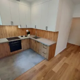 Nowoczesna kuchnia z białymi szafkami górnymi i drewnopodobnymi szafkami dolnymi, szarym blatem i płytkami podłogowymi, z widokiem na drewnianą podłogę w sąsiednim pomieszczeniu.