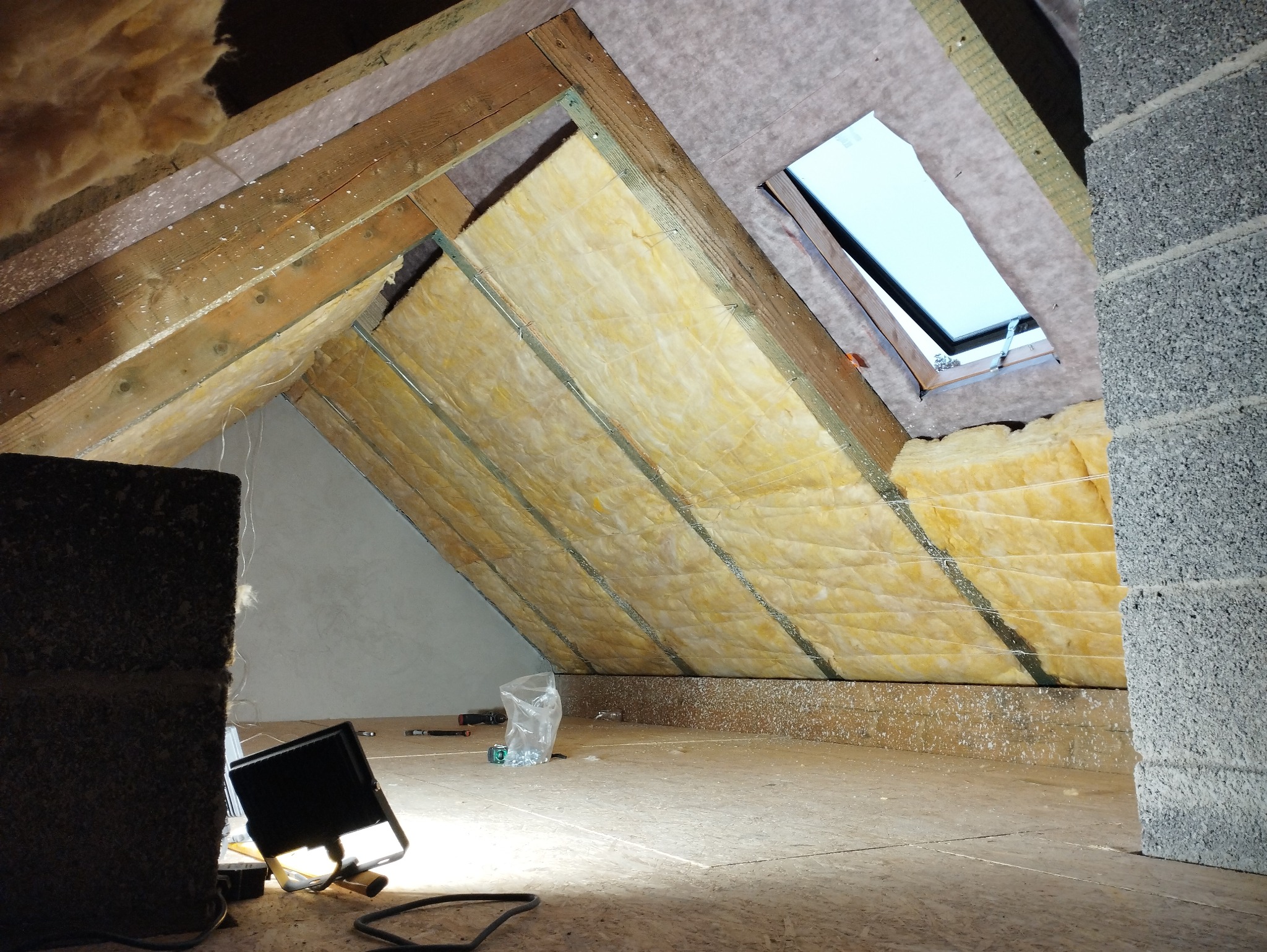 Ocieplenie poddasza wełną mineralną, widoczne elementy konstrukcji drewnianej, okno dachowe, fragment ściany z bloczków betonowych oraz lampa robocza.