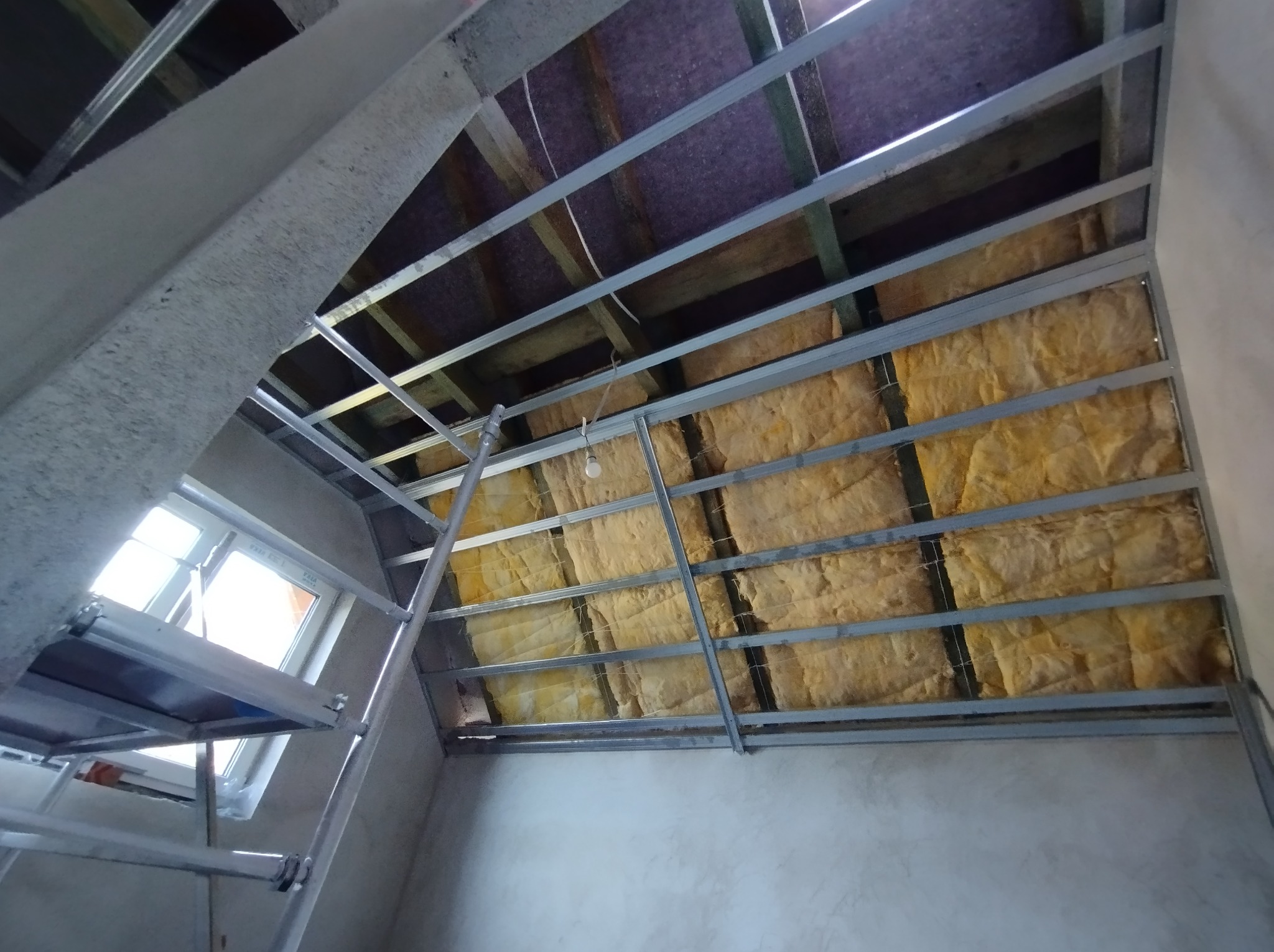 Montaż izolacji z wełny mineralnej w metalowym stelażu pod płytę gipsowo-kartonową na poddaszu, widoczny fragment okna dachowego i rusztowania.