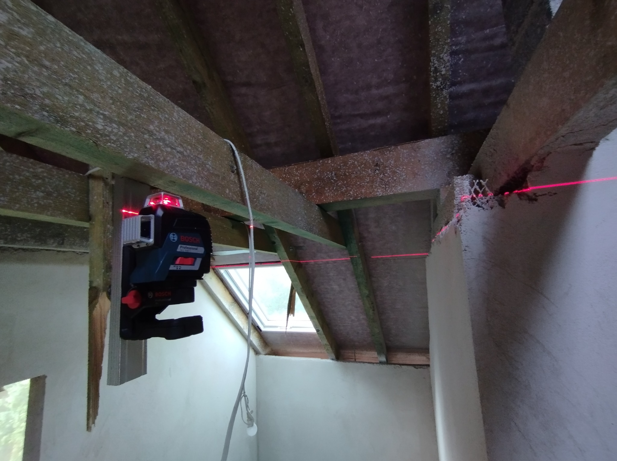 Laser krzyżowy Bosch ustawiony na drewnianej belce, wyznaczający poziom na ścianie poddasza w trakcie prac wykończeniowych.