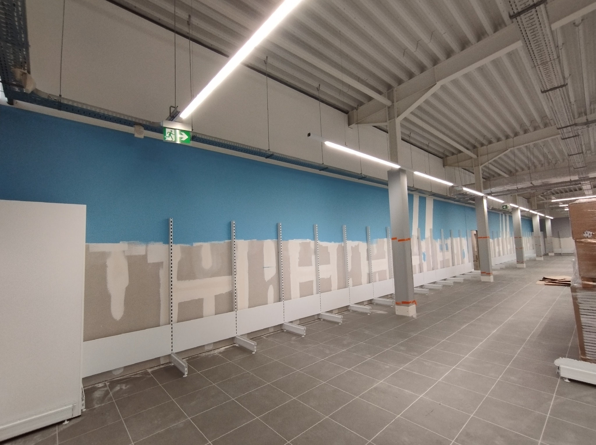 Przygotowanie ścian pod regały w nowym lokalu handlowym: częściowo pomalowana ściana na niebiesko, widoczne fragmenty szpachli i montowane metalowe wsporniki pod półki.