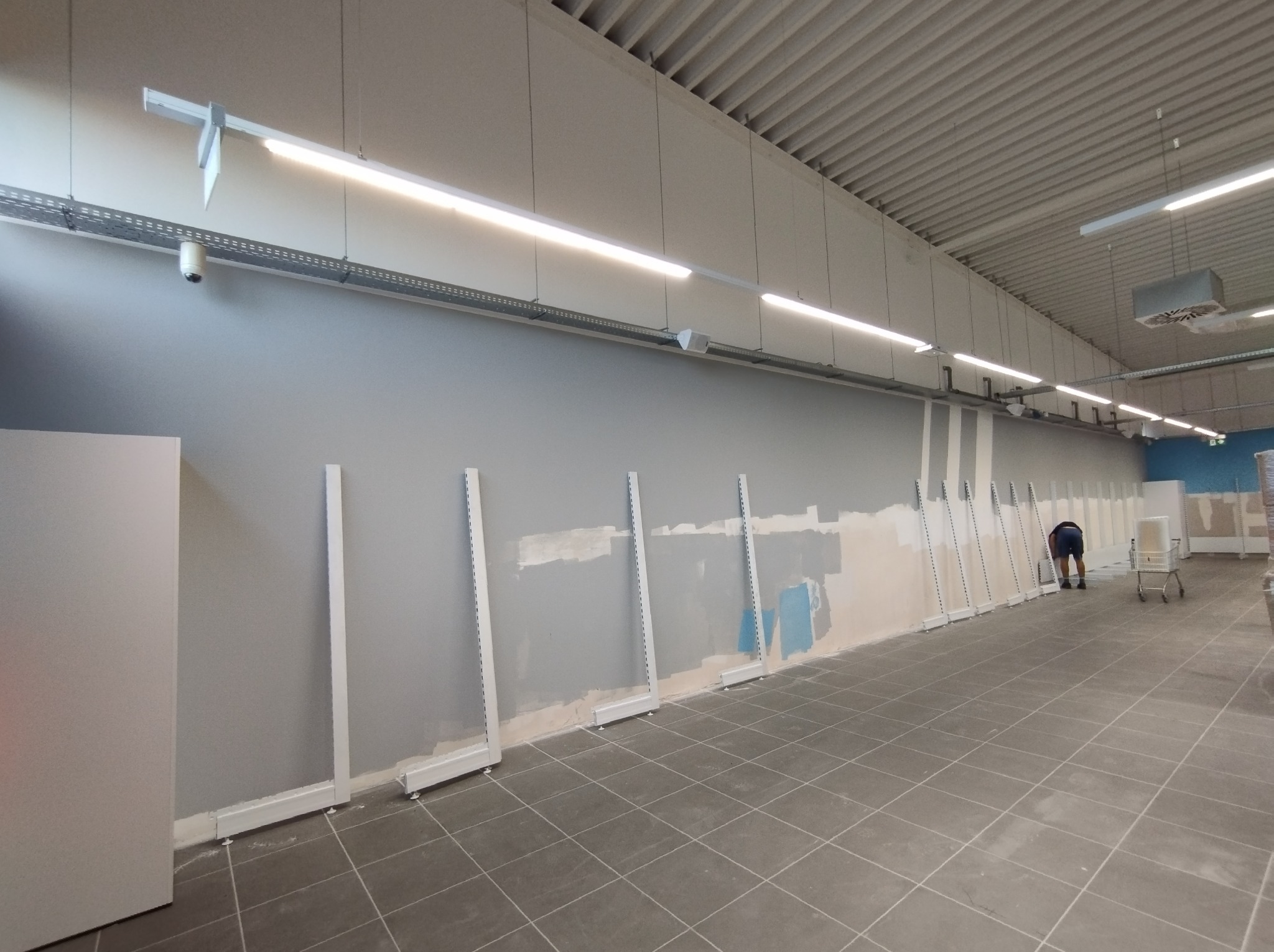 Przygotowanie ściany w dużym pomieszczeniu do montażu regałów: szpachlowanie i malowanie na szaro, widoczne białe wsporniki, osoba w tle z wózkiem.