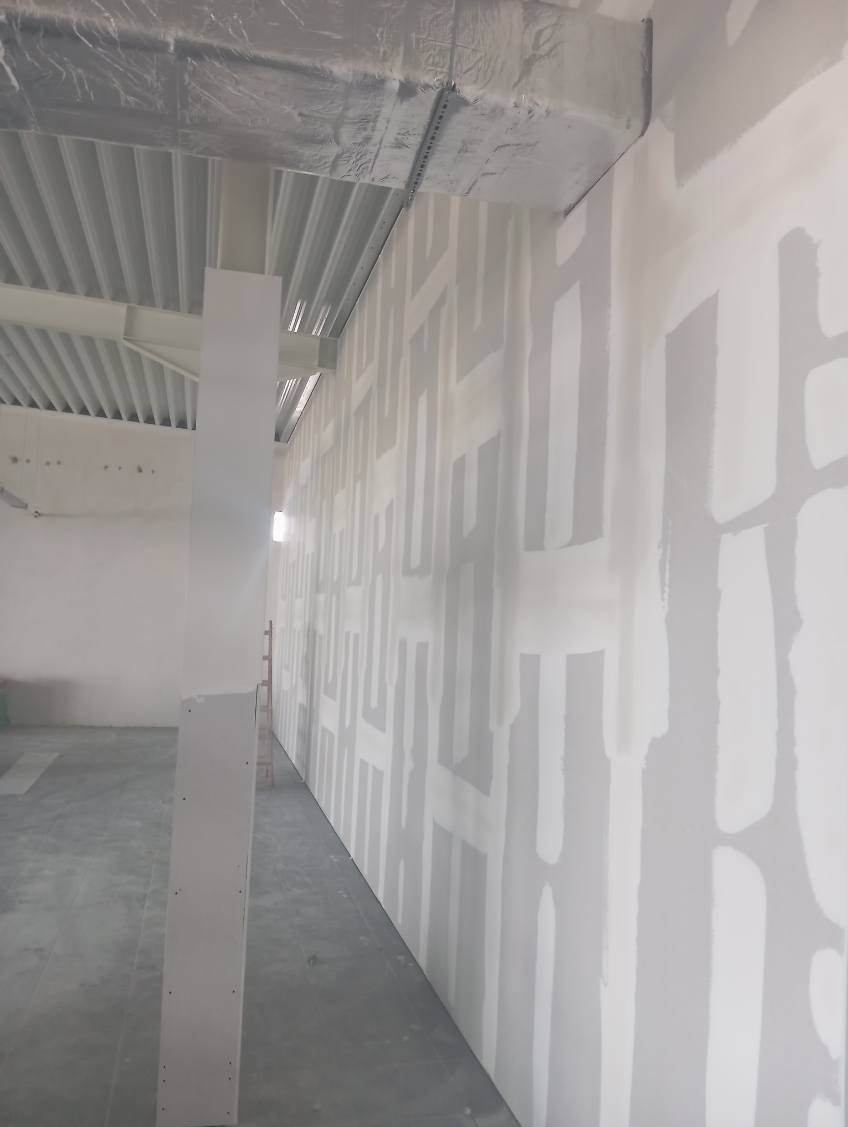 Wnętrze w trakcie prac wykończeniowych: ściana z płyt kartonowo-gipsowych, częściowo zaszpachlowana, widoczna metalowa konstrukcja sufitu i betonowa posadzka.