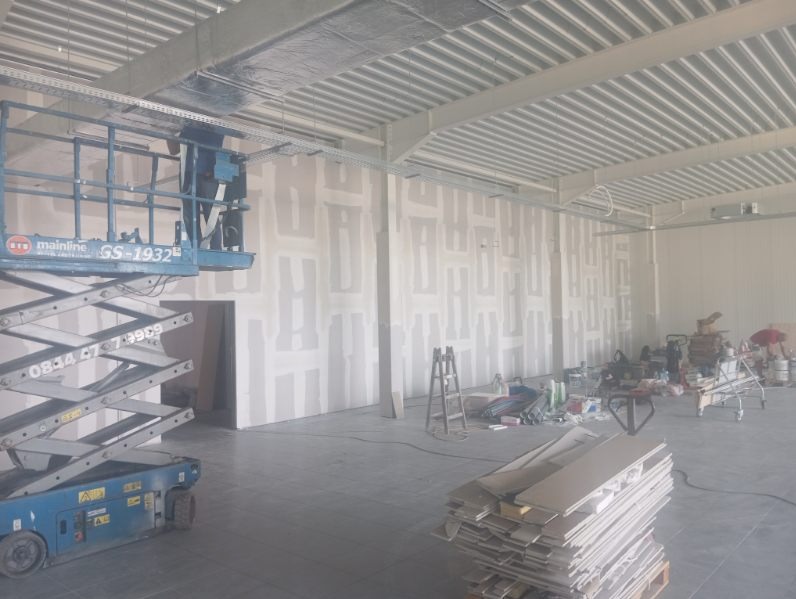 Wnętrze hali w trakcie prac wykończeniowych, widoczne płyty kartonowo-gipsowe na ścianach przed szpachlowaniem, podnośnik koszowy po lewej stronie oraz materiały budowlane na podłodze.