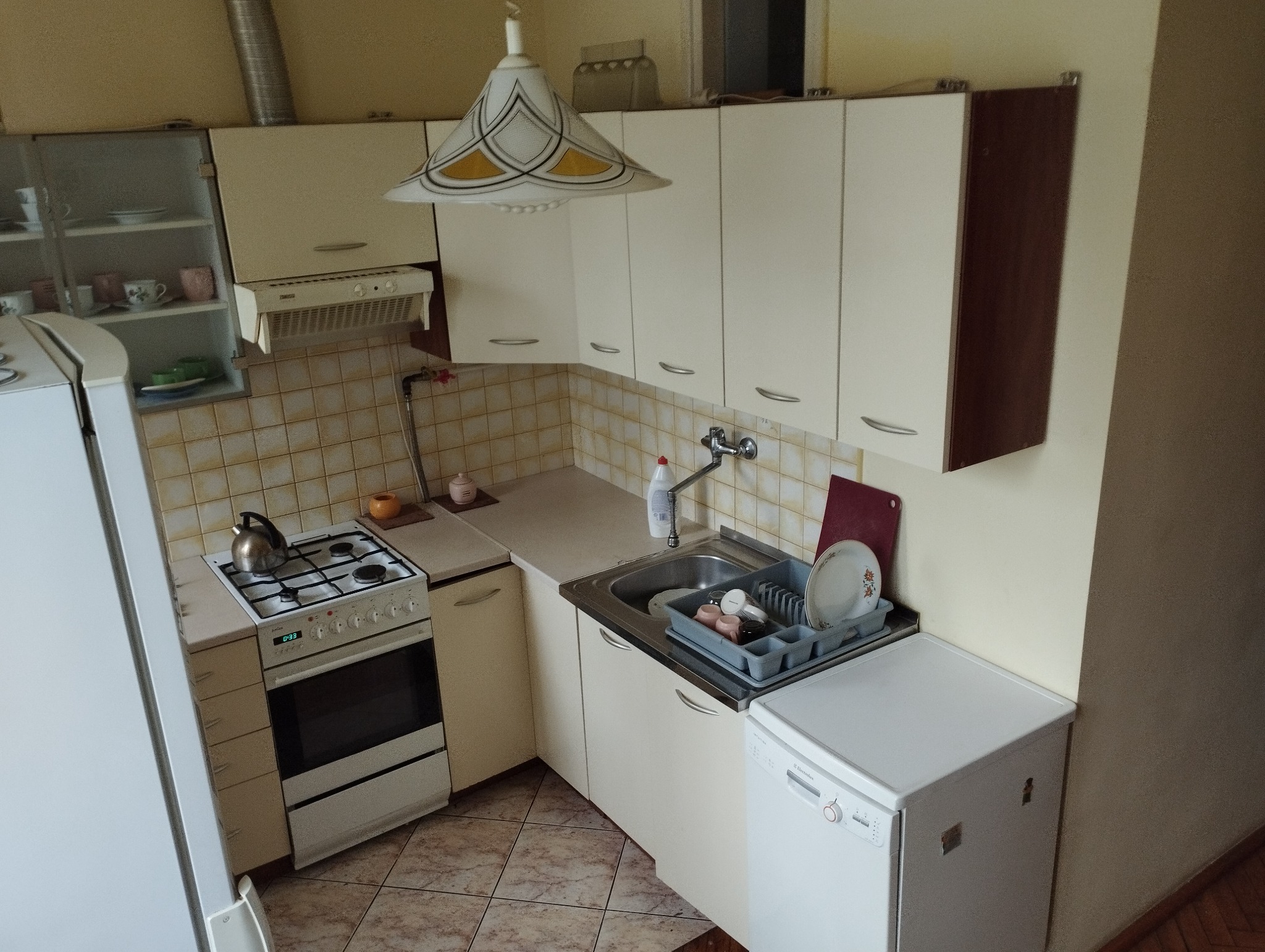 Kuchnia z beżowymi szafkami, płytkami i brązowymi akcentami, widok na zlew z naczyniami w suszarce, kuchenkę z czajnikiem i białą lodówkę.