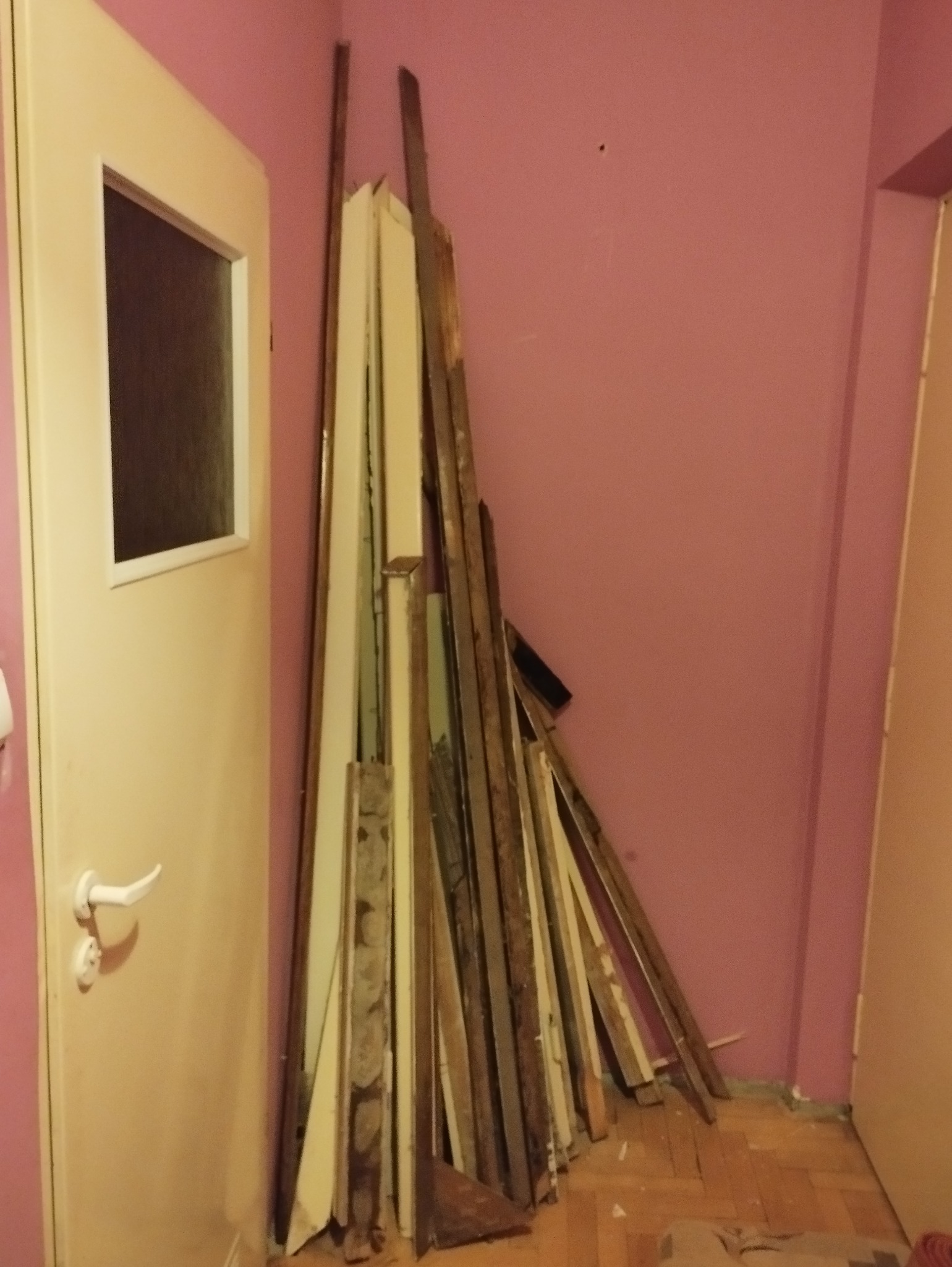 Stos starych, drewnianych listew przypodłogowych i elementów wykończeniowych oparty o ścianę w pokoju z drzwiami z szybą. Widoczne ślady demontażu i przygotowania do dalszych prac.