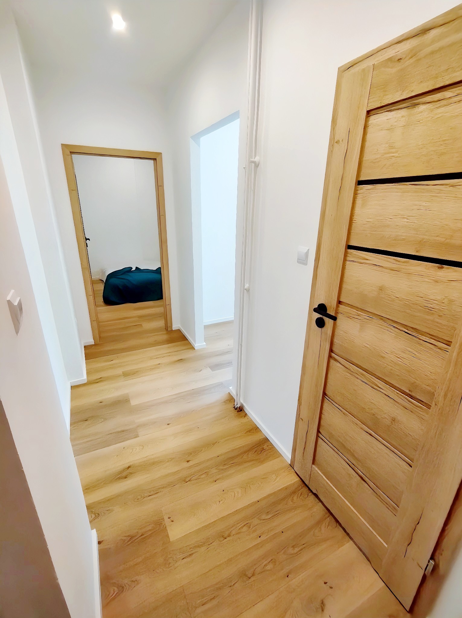 Jasny korytarz z drewnianą podłogą i drzwiami o naturalnym wykończeniu, widok na otwarte przejście do innego pokoju z widocznym łóżkiem.