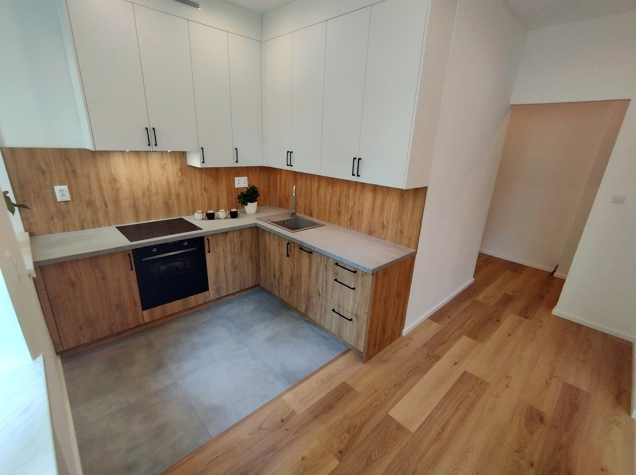 Nowoczesna kuchnia z białymi szafkami górnymi i drewnopodobnymi szafkami dolnymi, szarym blatem i płytkami podłogowymi, z widokiem na drewnianą podłogę w sąsiednim pomieszczeniu.