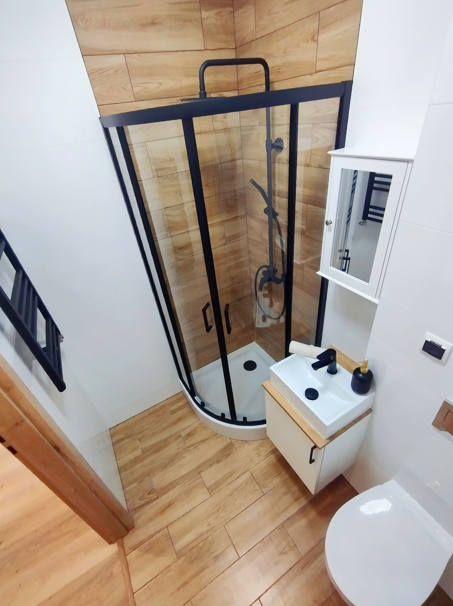 Nowoczesna łazienka z kabiną prysznicową z czarnymi ramami, umywalką nablatową z czarną armaturą i drewnopodobnymi płytkami na ścianach i podłodze.