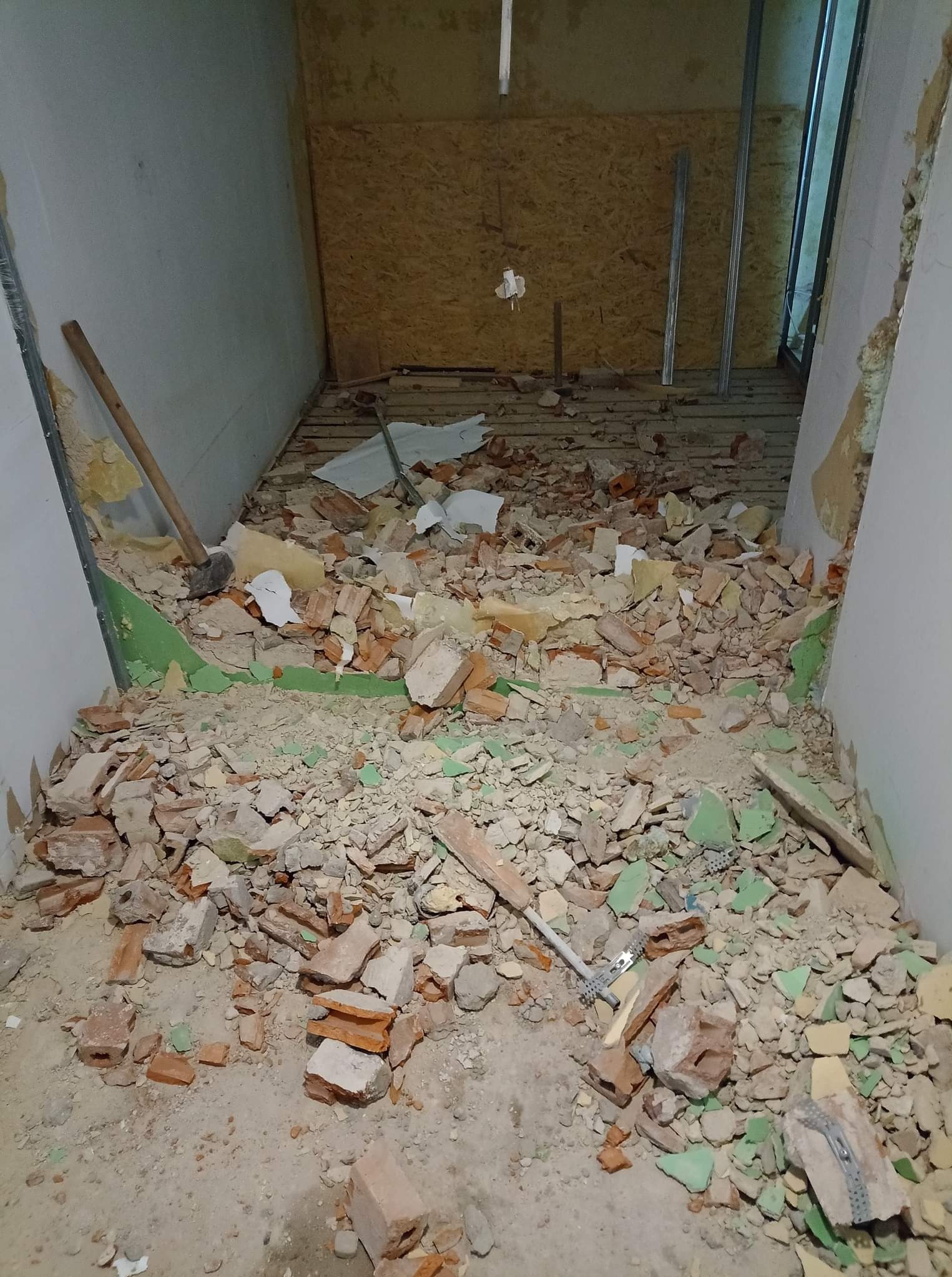 Wnętrze po rozbiórce: gruz ceglany i fragmenty płyt karton-gips z elementami metalowymi na podłodze w pomieszczeniu z fragmentami odsłoniętej boazerii i drewnianej ściany.