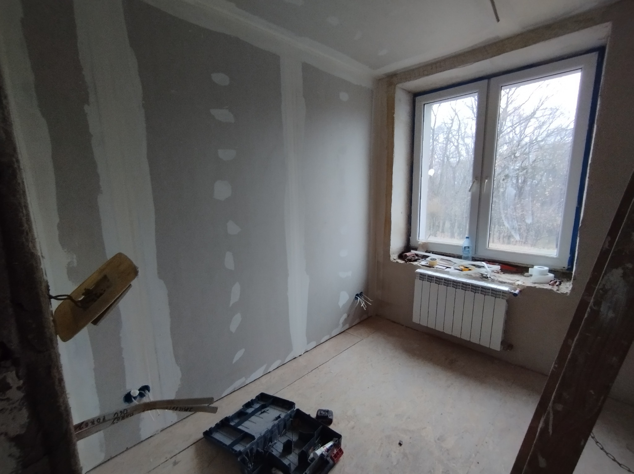 Ściana z płyt karton gipsowych w trakcie szpachlowania, widoczne narzędzia i skrzynka narzędziowa w pomieszczeniu w Tuszowie Narodowym, okno z widokiem na drzewa.