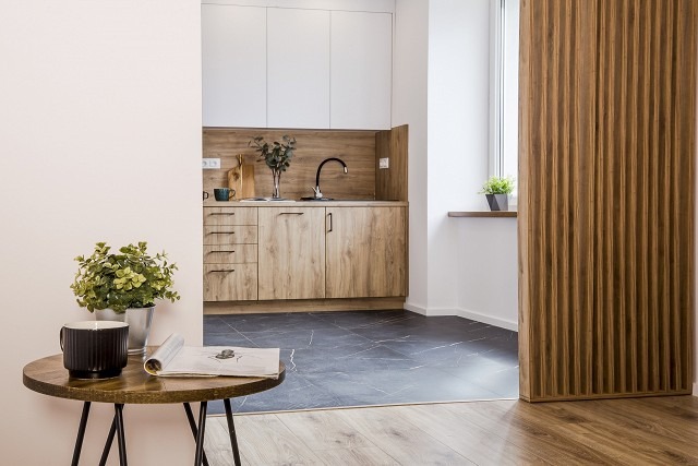 Nowoczesna kuchnia z drewnianymi szafkami, czarnym blatem i marmurową podłogą, widok z salonu z okrągłym stolikiem kawowym i drewnianą ścianką działową.