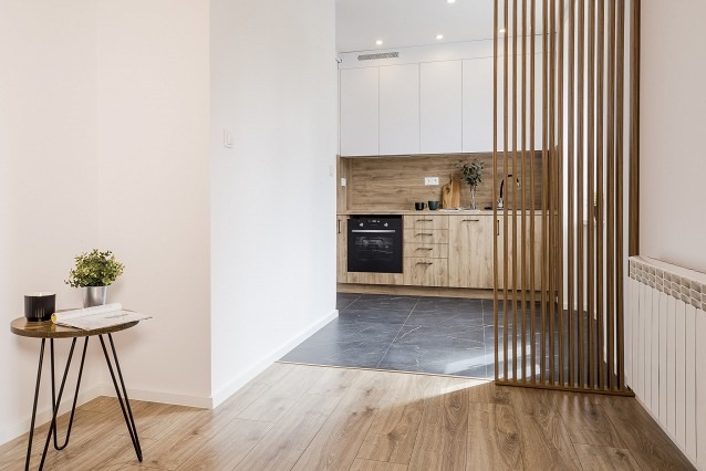 Jasne wnętrze mieszkania z drewnianą podłogą, stolikiem z rośliną i widokiem na kuchnię z drewnianymi szafkami i czarnym piekarnikiem, oddzieloną ażurową ścianką.