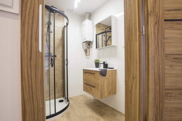 Nowoczesna łazienka z kabiną prysznicową z czarnymi profilami, wiszącą szafką z drewnianym fornirem, białymi płytkami ściennymi i grzejnikiem gazowym.