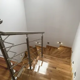 Widok z góry na drewniane schody z metalową balustradą, oświetlone małymi lampkami ściennymi, z widocznym odbiciem światła na lakierowanej powierzchni stopni i wiszącą żarówką na suficie.