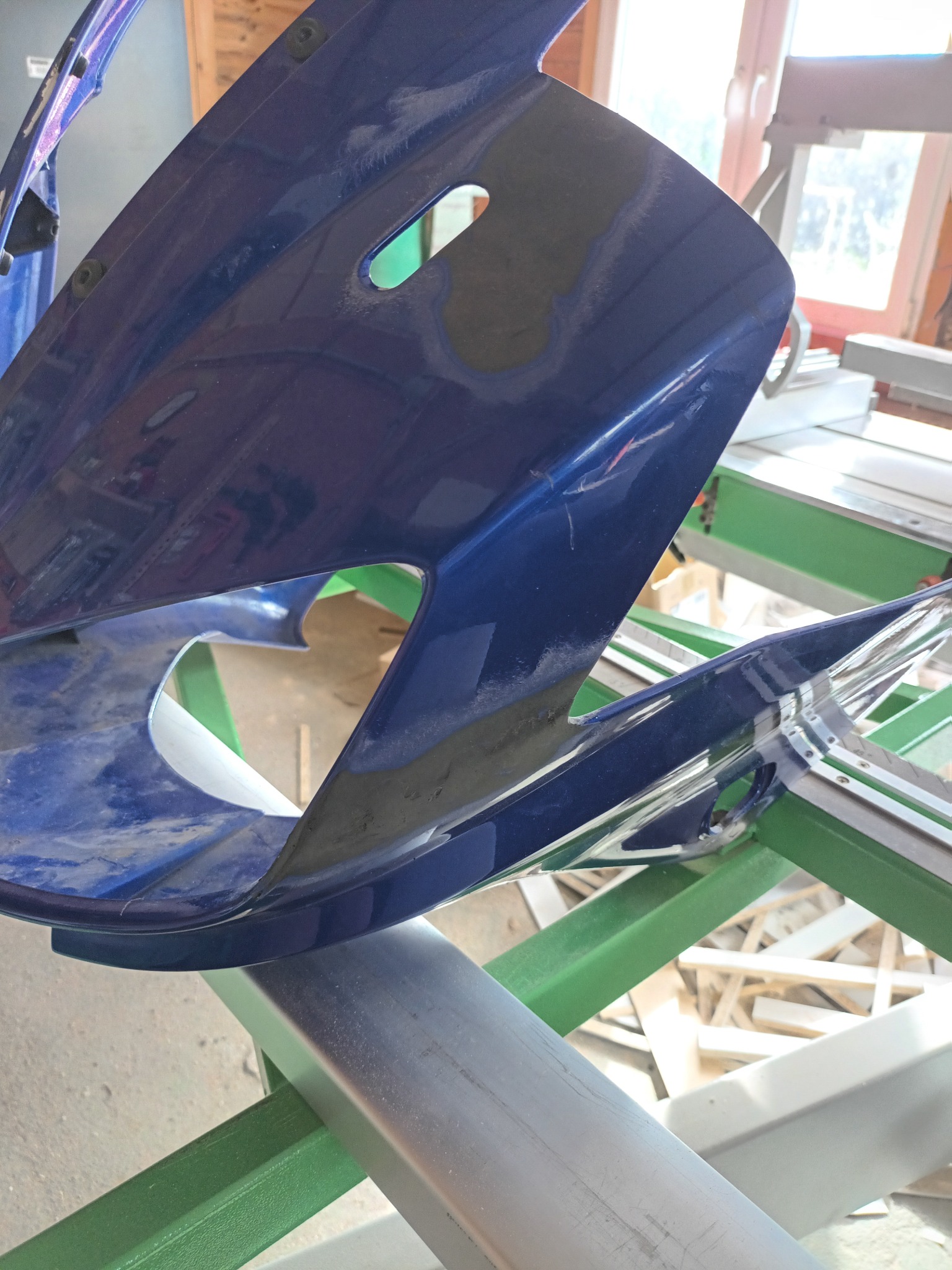 Niebieski element karoserii motocykla w trakcie naprawy, widoczne szpachlowane obszary, zamocowany na zielonym metalowym stelażu.