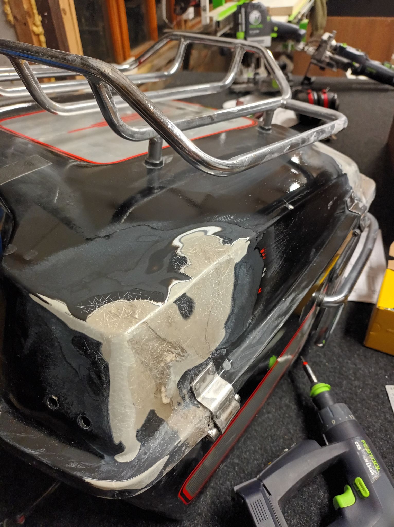 Czarny kufer motocyklowy z metalowym stelażem w trakcie naprawy, z widocznymi śladami szlifowania i nałożonym szpachlem, obok leży wkrętarka akumulatorowa.
