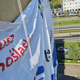 Na Poziomie Prace Alpinistyczne Tomasz Drapikowski - Pracownik w uprzęży zawieszony na linie montuje duży baner reklamowy na ścianie budynku, widok z góry na ulicę i budynki w tle.