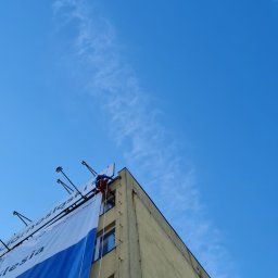 Na Poziomie Prace Alpinistyczne Tomasz Drapikowski - Pracownik w czerwonym kombinezonie zabezpieczony linami, montuje duży baner reklamowy na ścianie budynku. Widok z dołu, na tle błękitnego nieba.