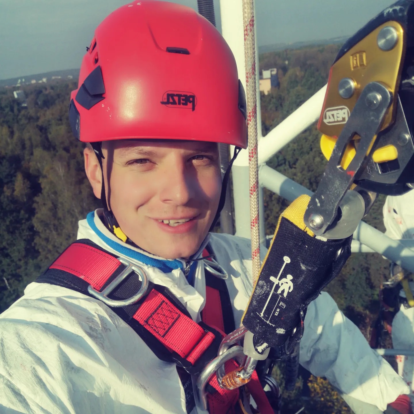 Uśmiechnięty pracownik wysokościowy w czerwonym kasku Petzl i białym kombinezonie, zabezpieczony uprzężą i sprzętem do zjazdu na tle konstrukcji i zieleni.