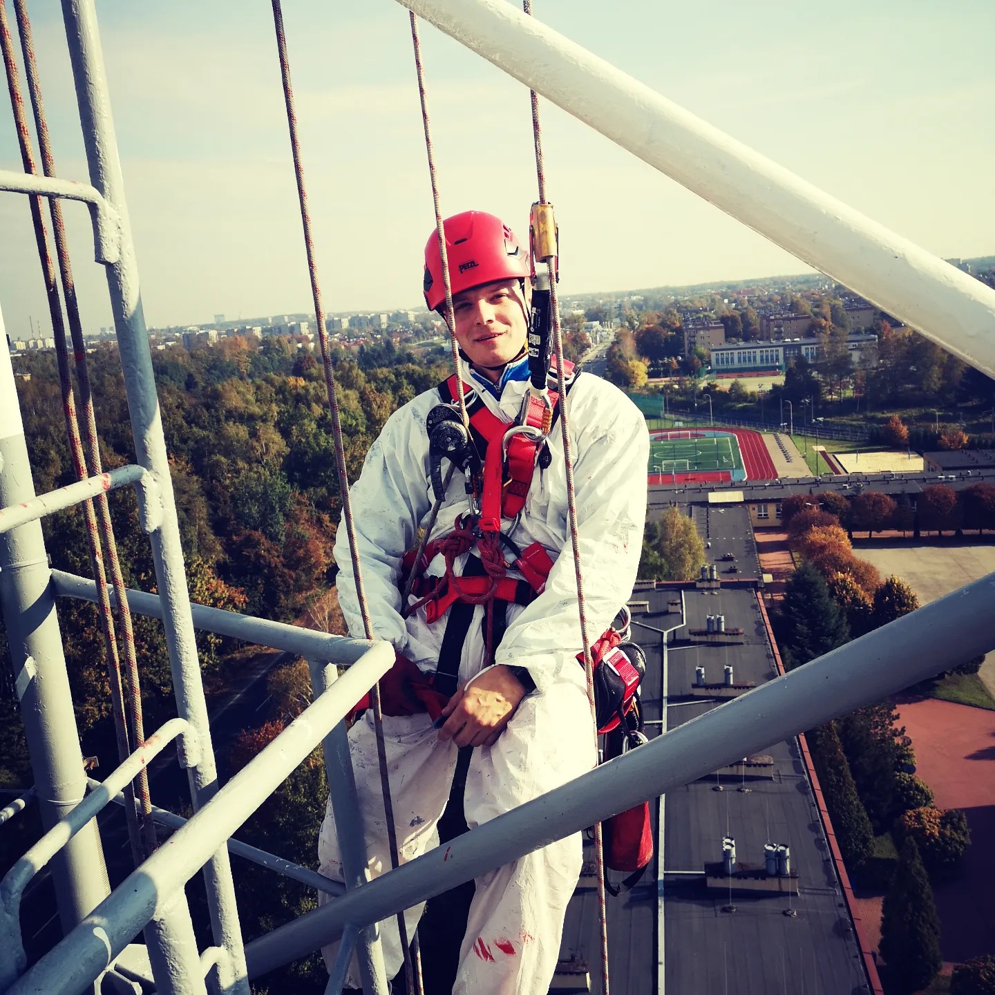 Mężczyzna w czerwonym kasku i białym kombinezonie, zabezpieczony linami, stoi na konstrukcji stalowej na wysokości, z widokiem na miasto z drzewami i stadionem w tle.