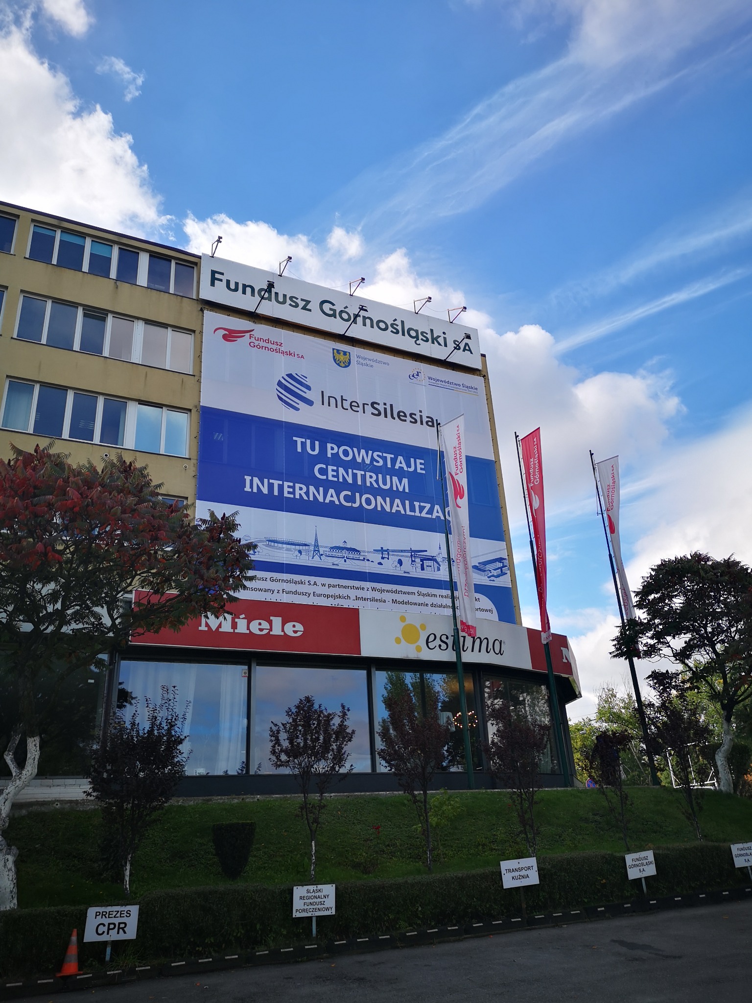 Budynek w Czechowicach-Dziedzicach z dużym banerem reklamowym Funduszu Górnośląskiego S.A. i flagami, widoczne drzewa i trawnik przed budynkiem.