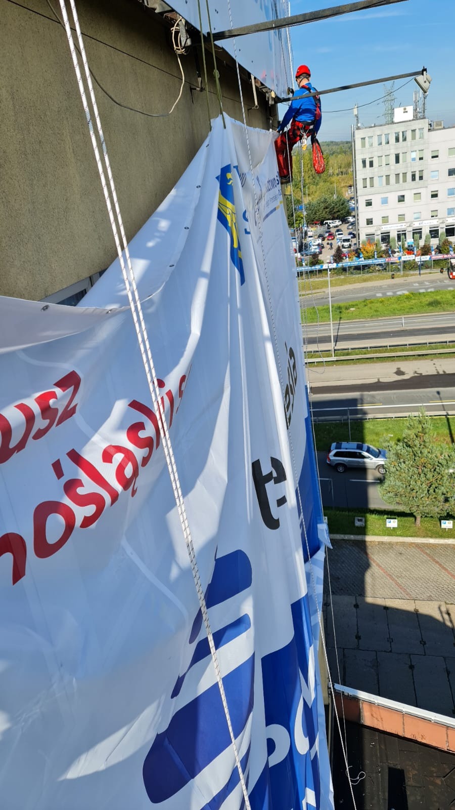 Pracownik w uprzęży zawieszony na linie montuje duży baner reklamowy na ścianie budynku, widok z góry na ulicę i budynki w tle.