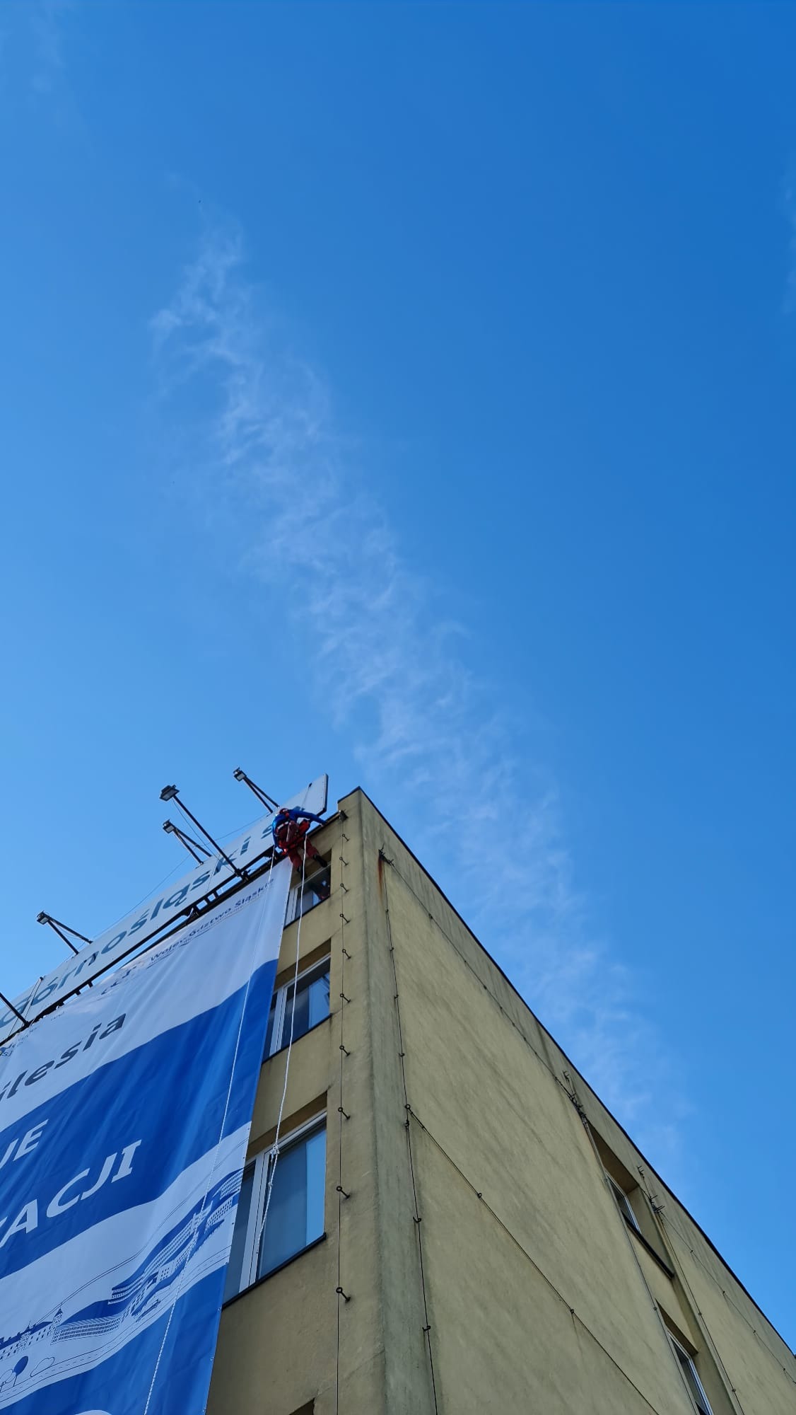 Pracownik w czerwonym kombinezonie zabezpieczony linami, montuje duży baner reklamowy na ścianie budynku. Widok z dołu, na tle błękitnego nieba.