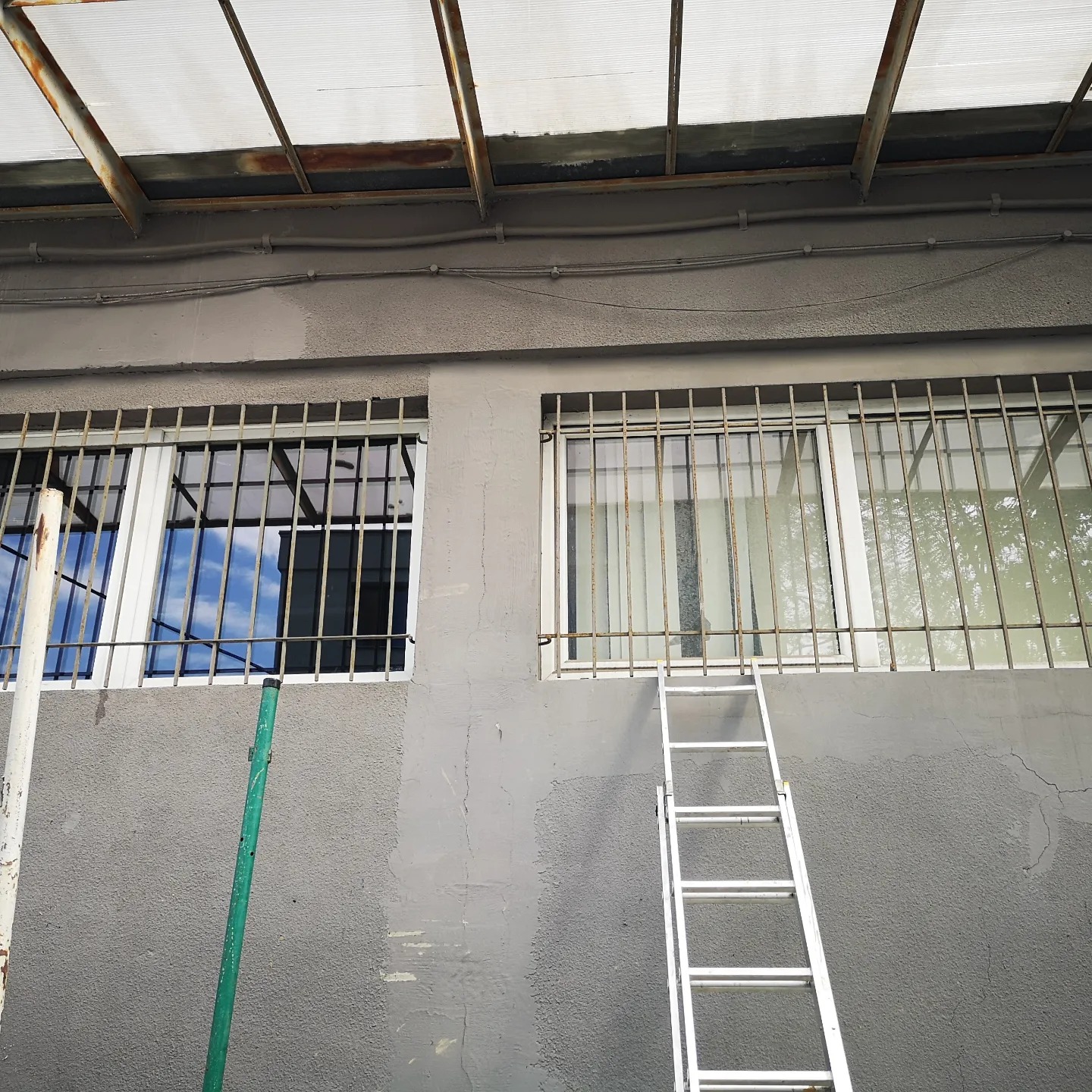 Szara ściana budynku z dwoma oknami zabezpieczonymi kratami, częściowo pomalowana, z opartą o nią drabiną i zielonym prętem.