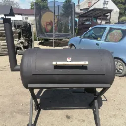 Czarny, spawany grill wędzarniczy z termometrem, ustawiony na zewnątrz na tle odwróconego samochodu, trampoliny i zaparkowanego auta.