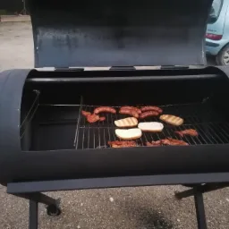 Czarny, metalowy grill beczka z otwartą pokrywą, na ruszcie kiełbaski i pieczywo, w tle zaparkowany samochód.