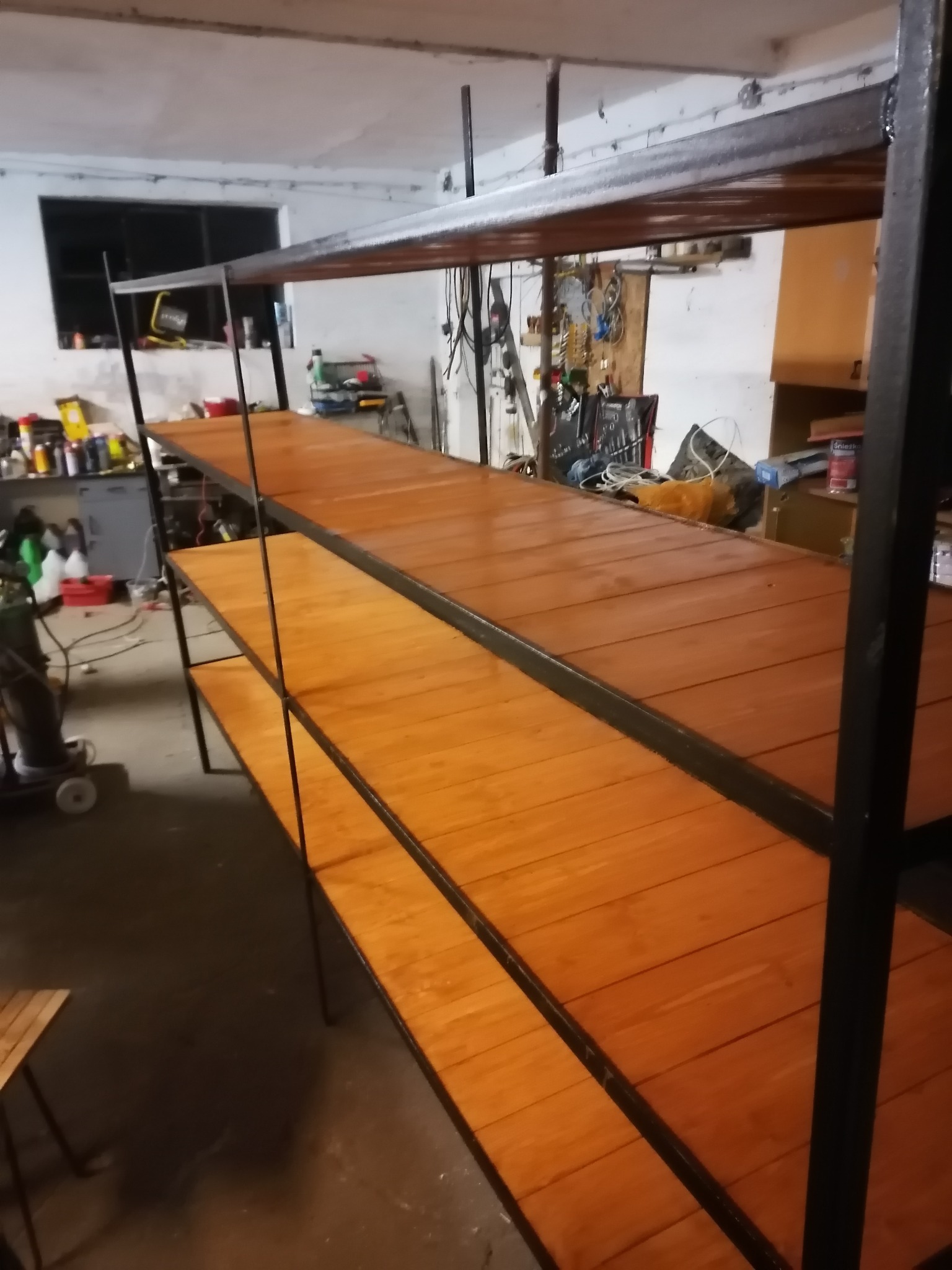 Metalowa konstrukcja regału z drewnianymi półkami w warsztacie, widoczne spawy i surowe wykończenie, oświetlenie warsztatowe.