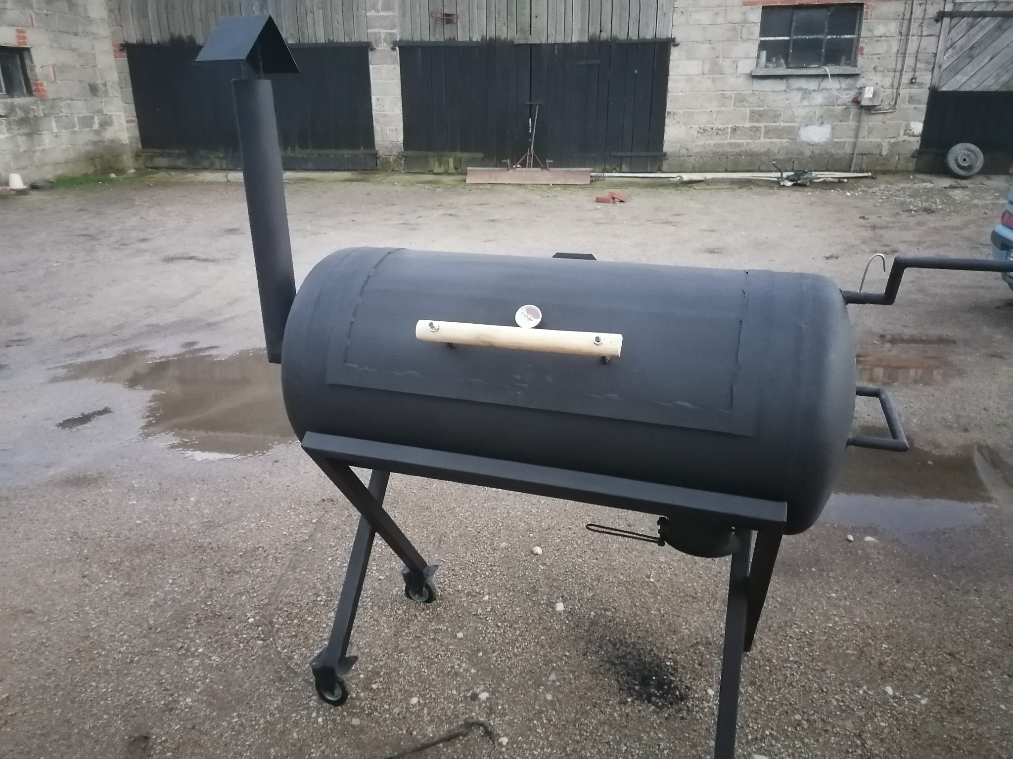Czarny, spawany grill w kształcie walca z drewnianą rączką i kominem, na zewnątrz, na tle murowanego budynku gospodarczego.